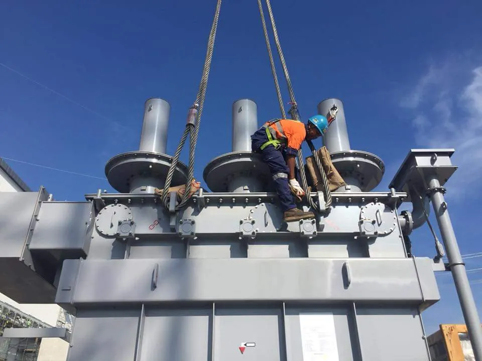 Rigger standing on top of a large transformer attached to crane hooks
